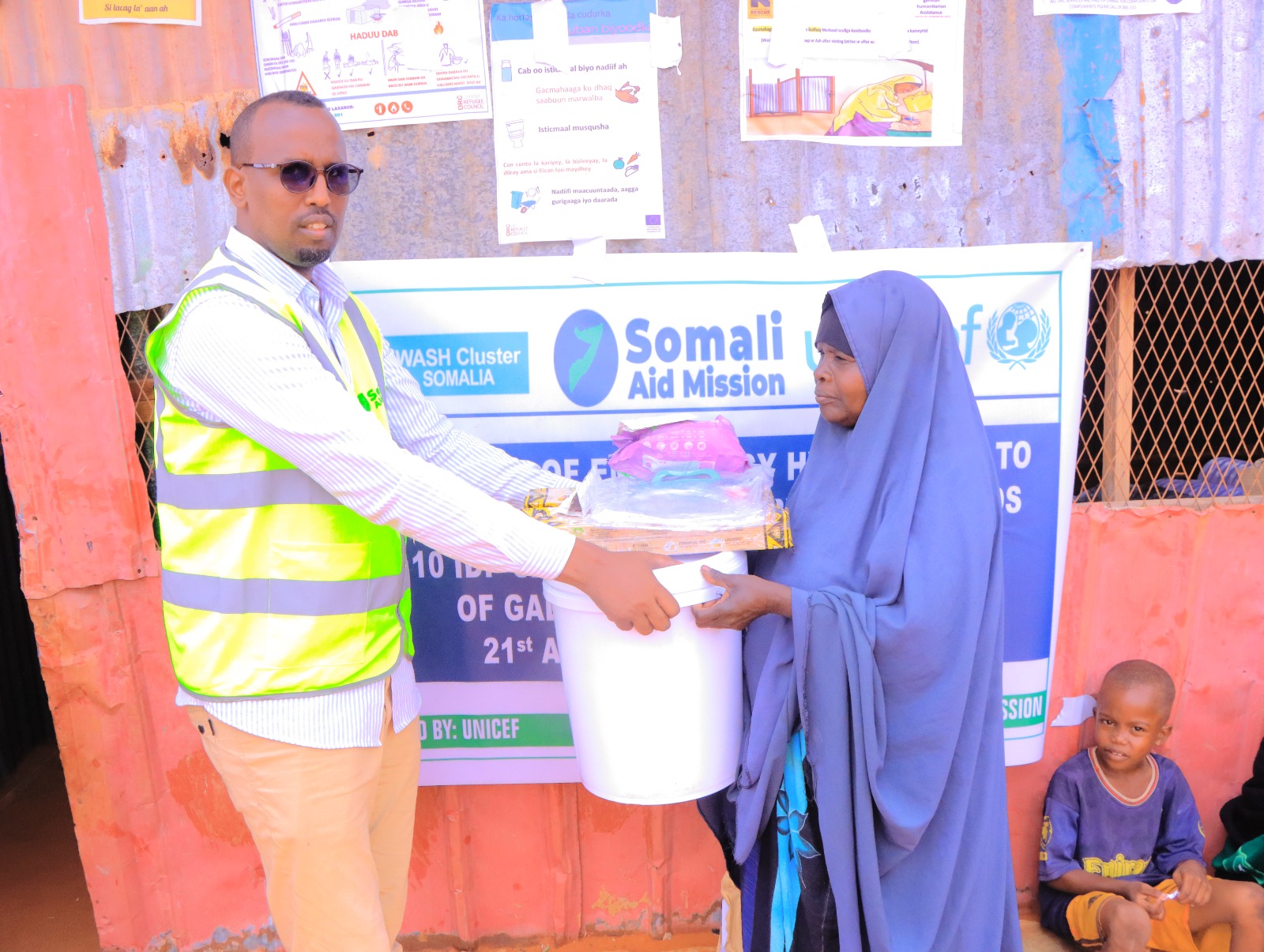 A Somali Aid Mission staff member hands a hygiene kit to a woman in front of a WASH Cluster and UNICEF banner